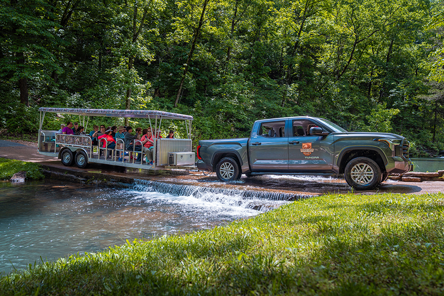 A pickup truck pulls a Dogwood Canyon tram car across a river crossing in the park. The tram is full of guests enjoying the sunshine.
