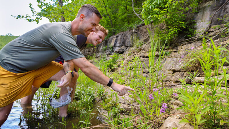 2025_DWC_Activities_CreekCrawlAdventure_Header_900x600 Educator and guest standing in the creek pointing at a something in the tall grass.